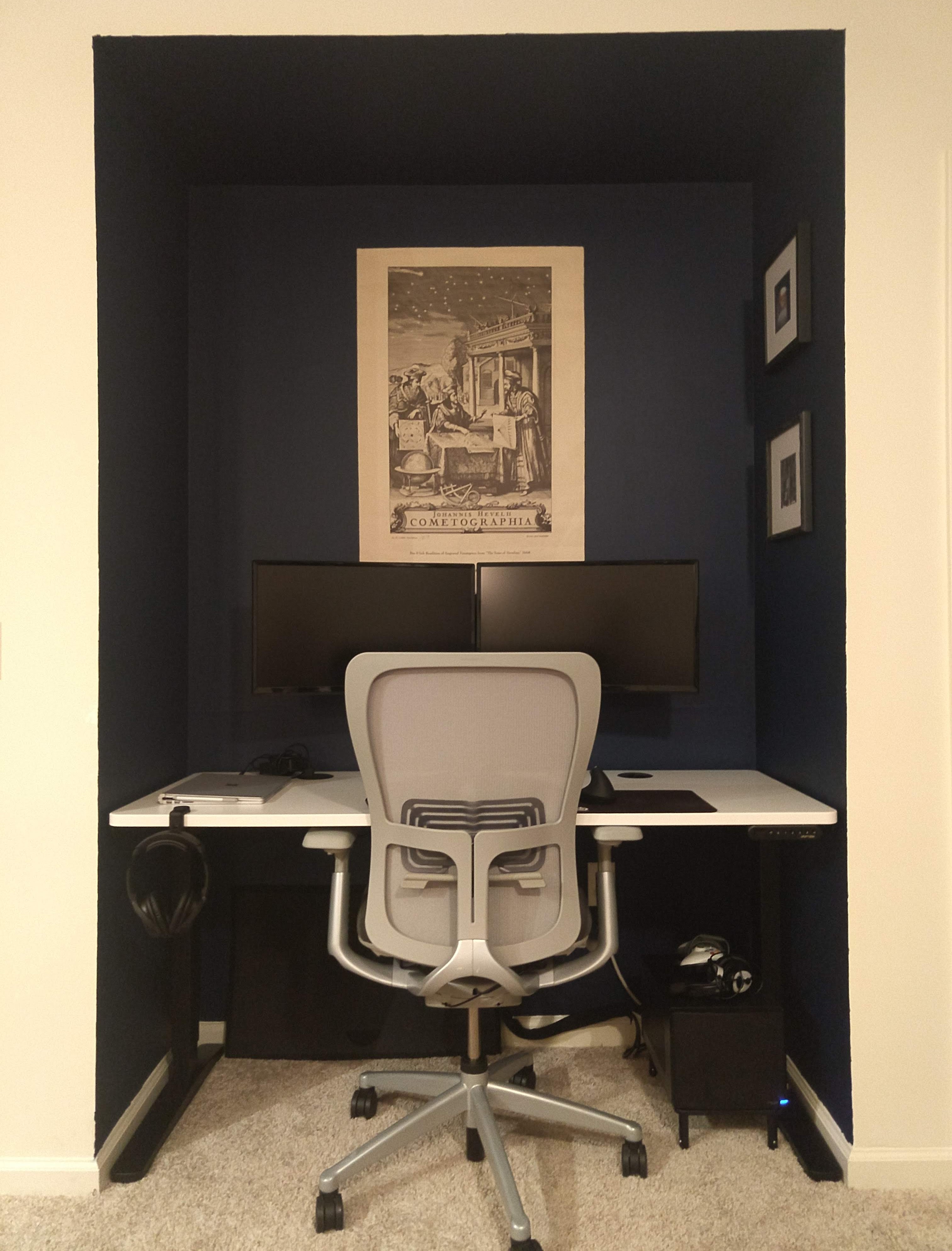 Photo of the final setup. A white desk surmounted by two monitors sits nestled in an alcove. The alcove is painted navy blue and has a poster and several framed photos hung up. A computer chair sits with its base tucked under the desk. A workstation sits on a platform above the floor on the bottom right.